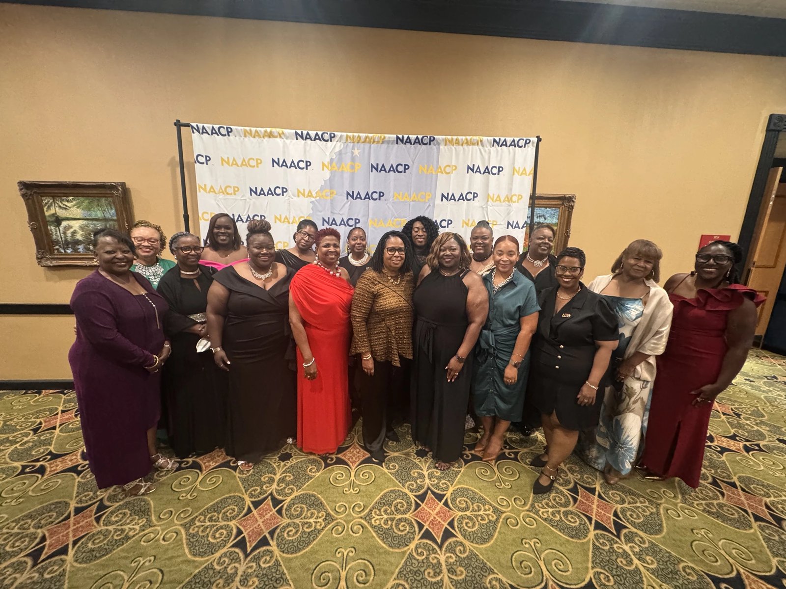 A group of women in formal dress posed in front of an NAACP step-and-repeat banner