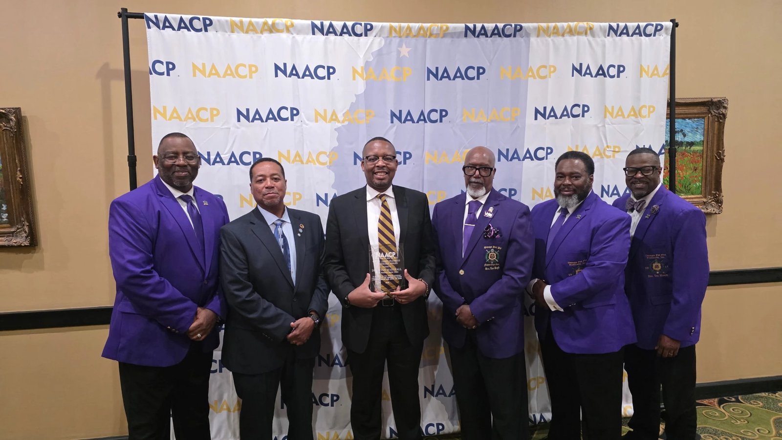 Dr. Jevon Marshall holding the Willie James Plunkett Leadership Award at the NAACP banner, surrounded by six men in the purple and gold of Omega Psi Phi Fraternity, Inc., including DeSoto County NAACP 1st Vice President Adrian Millsap